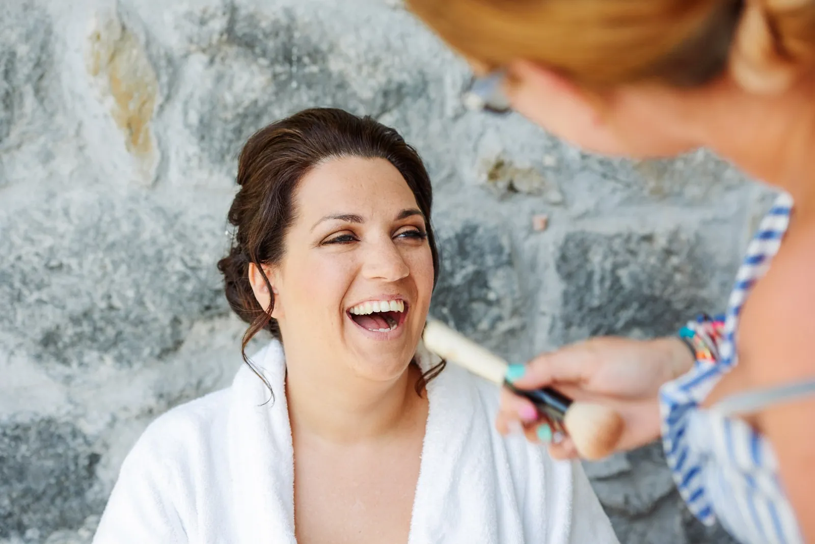 bride laughing whilst having her makeup applied in front of a grey stone wall