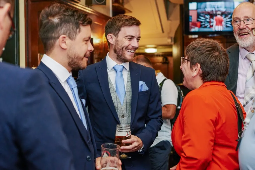 Groom laughing and drinking beer in a London pub with wedding guests