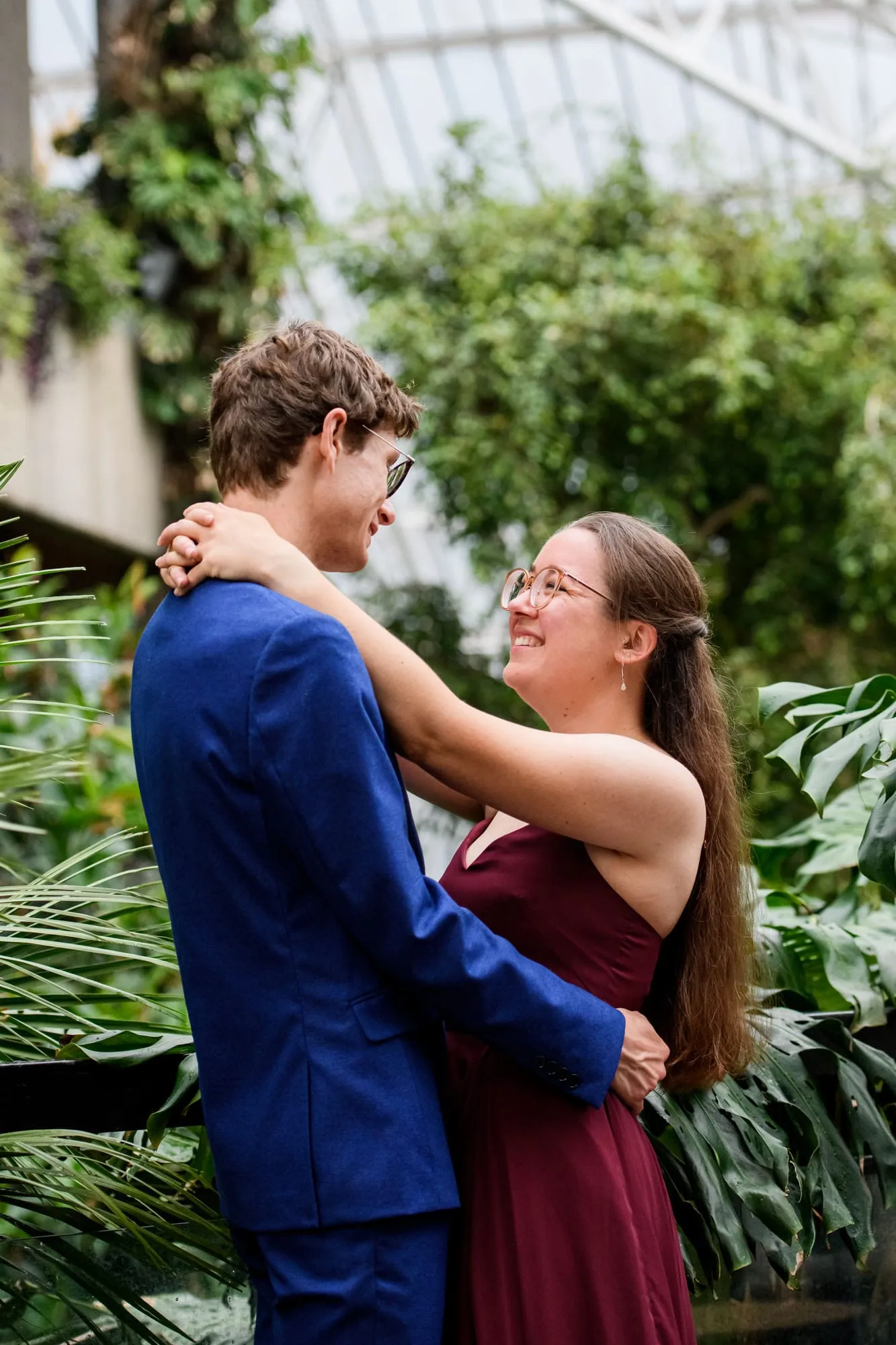 Engaged couple smiling with arms around each other whilst surrounded by plants