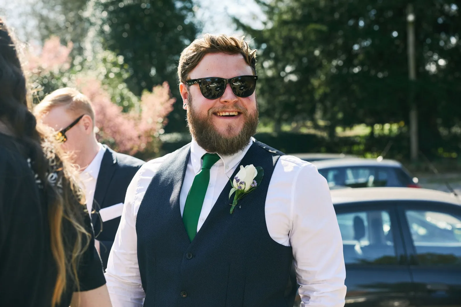 Natural wedding guest smiling whilst stood outside in a blue waistcoat