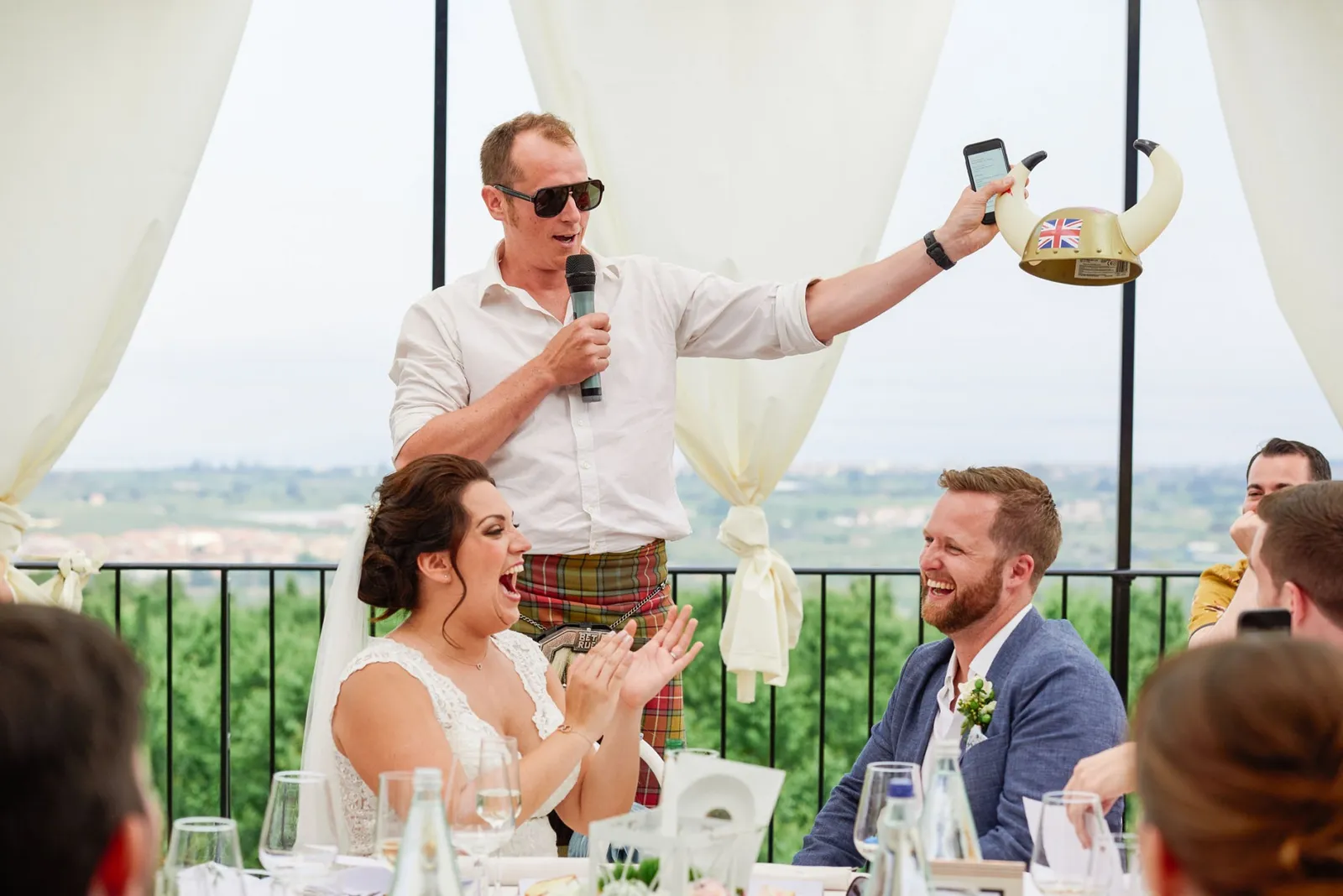 Best man giving a speech whilst bride and groom laugh on their wedding day. with a scenic view of Sicily behind them