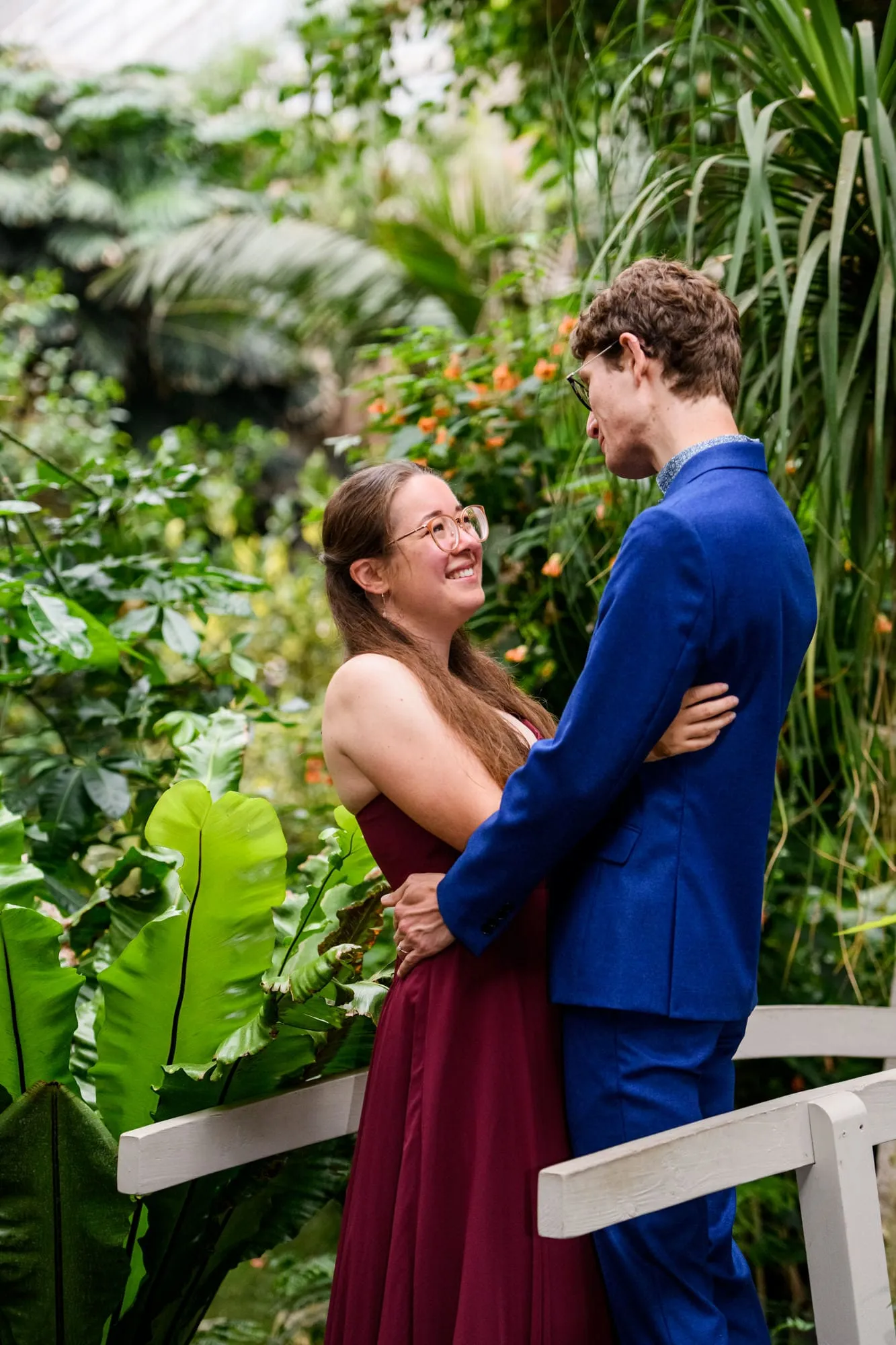 Engaged couple smiling with arms around each other whilst surrounded by plants
