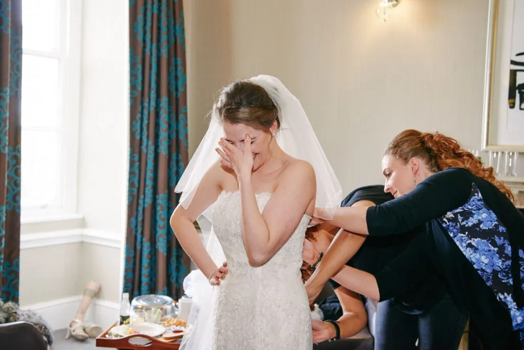 bride laughing whilst having her dress done up by friends