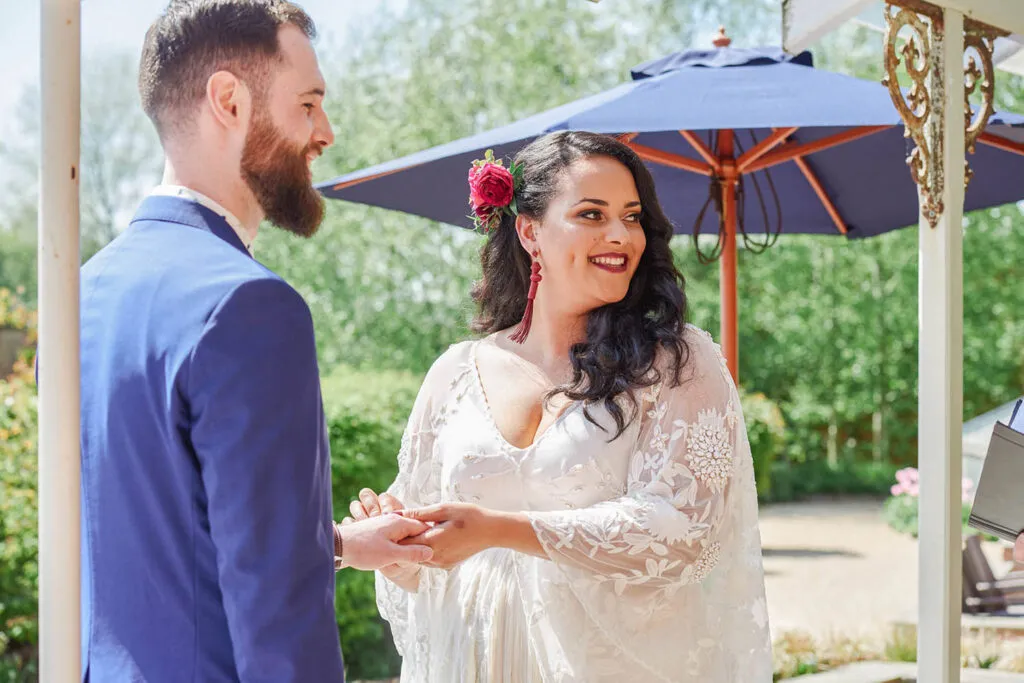 Bride smiling whilst putting ring on grooms hand during outdoor wedding in a garden