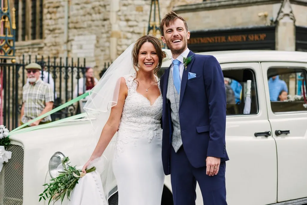 Bride and groom laughing together in front of white wedding car