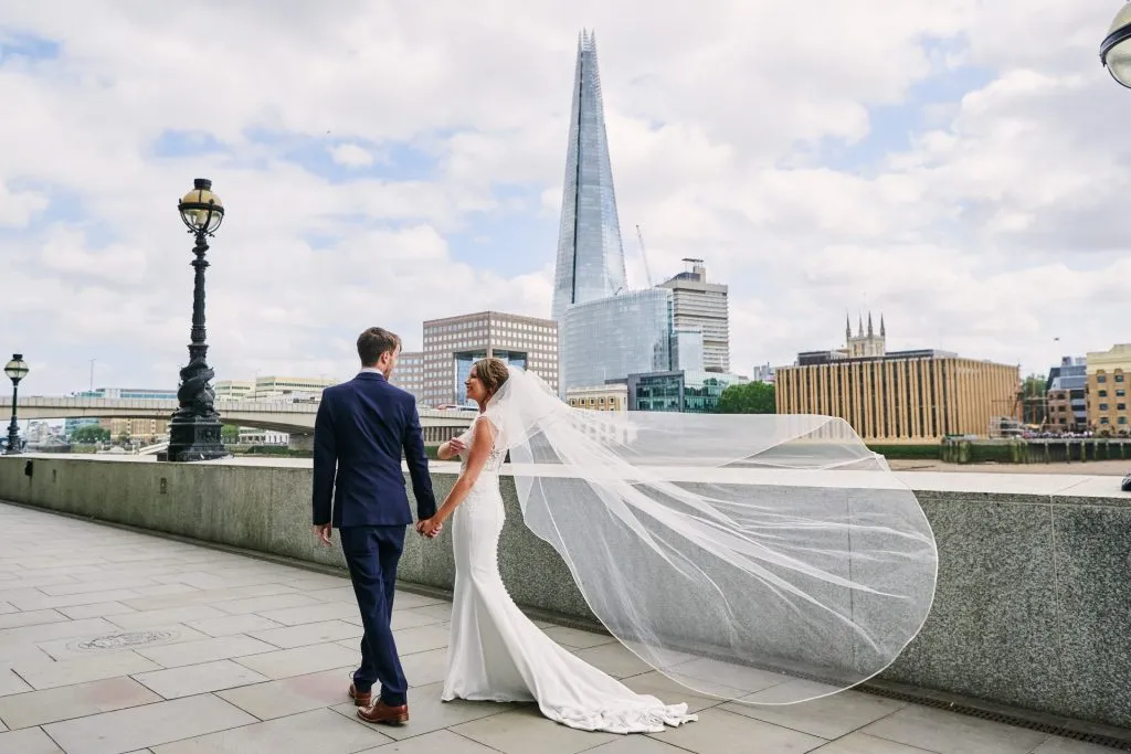 bride and groom walking along the riverside whilst veil blows in the wind