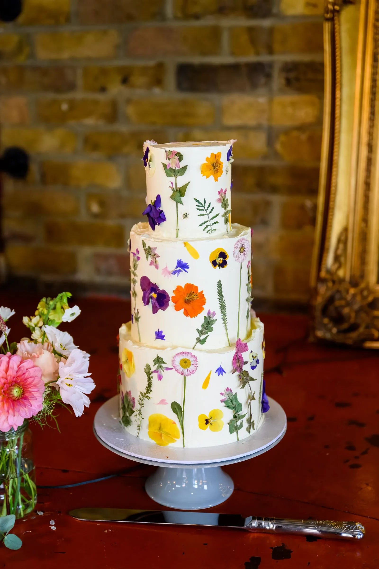 pressed flowers on white wedding cake in front of rustic brick wall at The Depot N7 