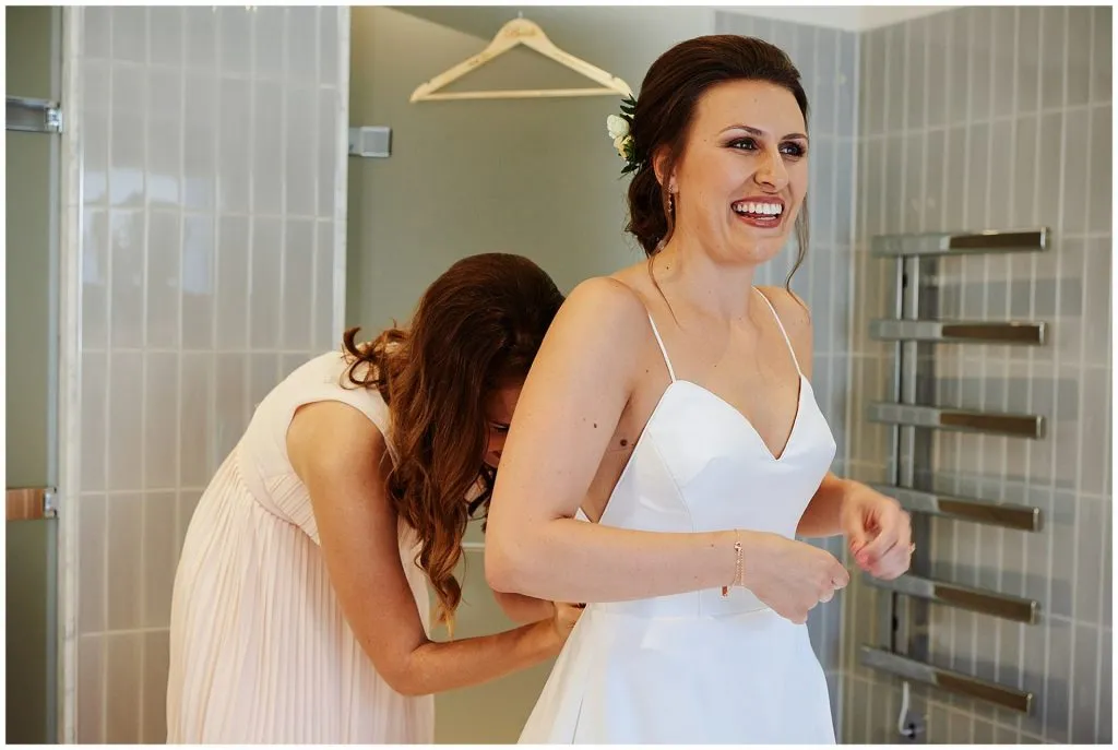 bride smiling whilst having her dress done up by her bridesmaid