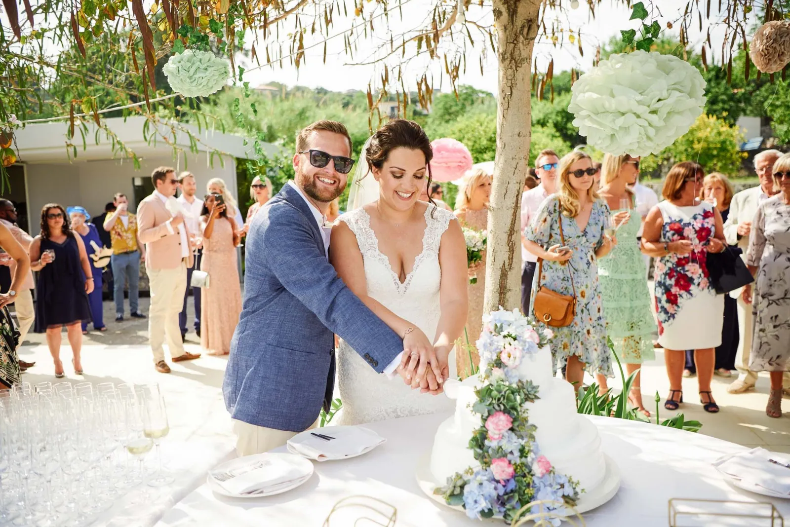 Bride and groom cutting their wedding cake whilst stood outside in Scilly surrounded by their guests.