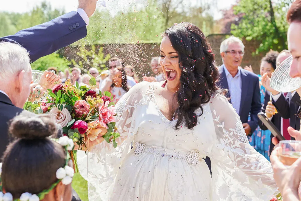 Bride celebrating as confetti is poured over her during outdoor summer wedding