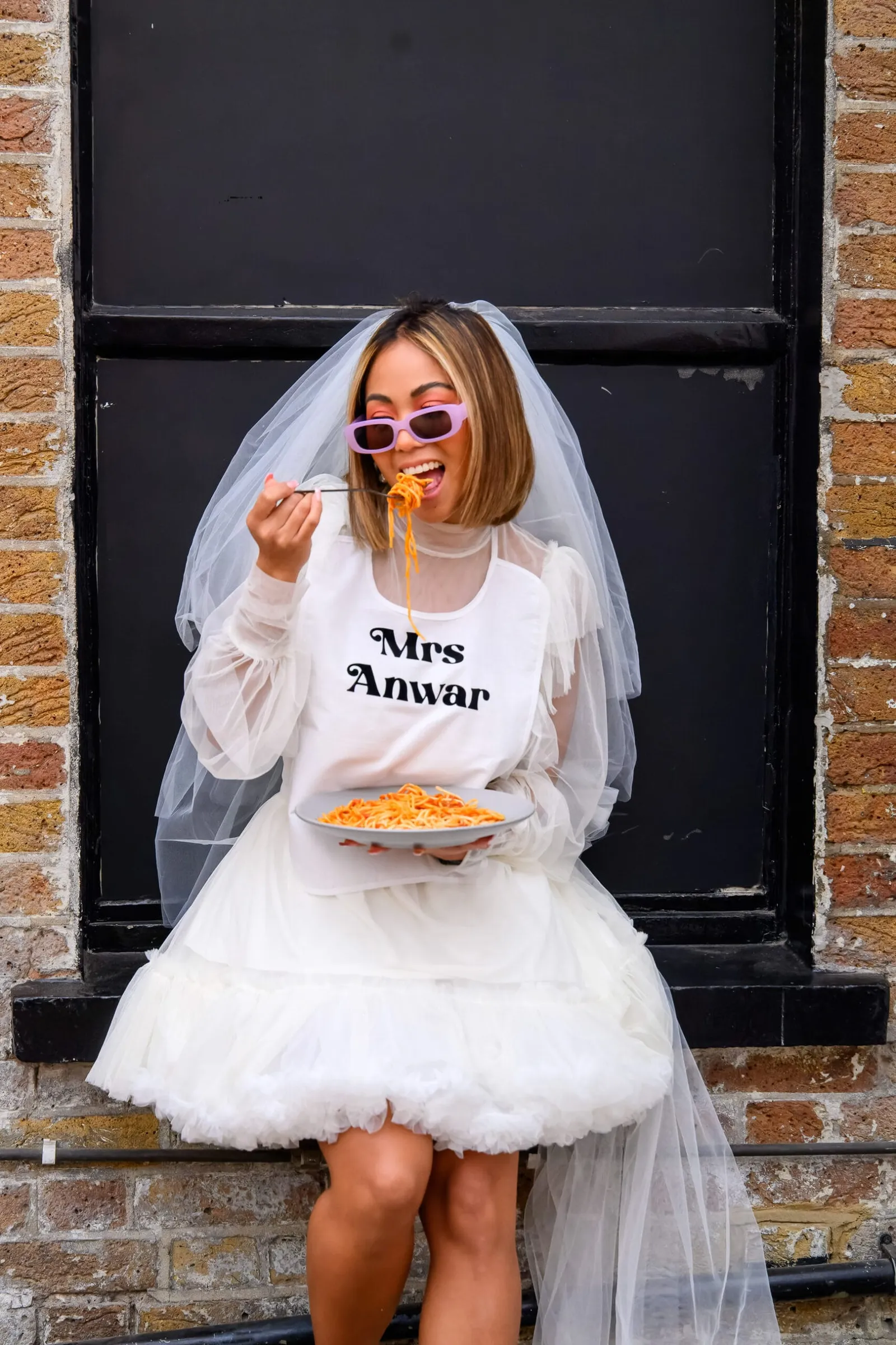 fun bride eating spaghetti sat on brick wall during wedding photos