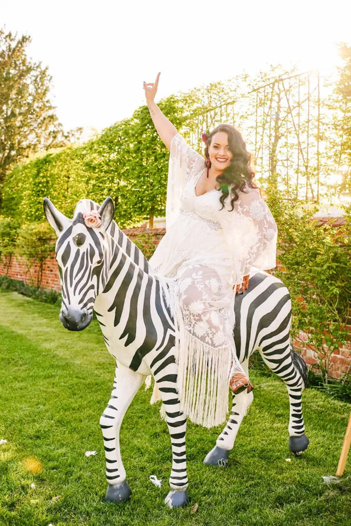 Fun bride sat smiling on a large prop zebra during Pippley Walled Garden summer wedding