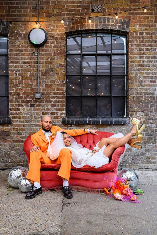 Fun and vibrant Nottingham wedding photography of stylish couple posing on red sofa against brick wall